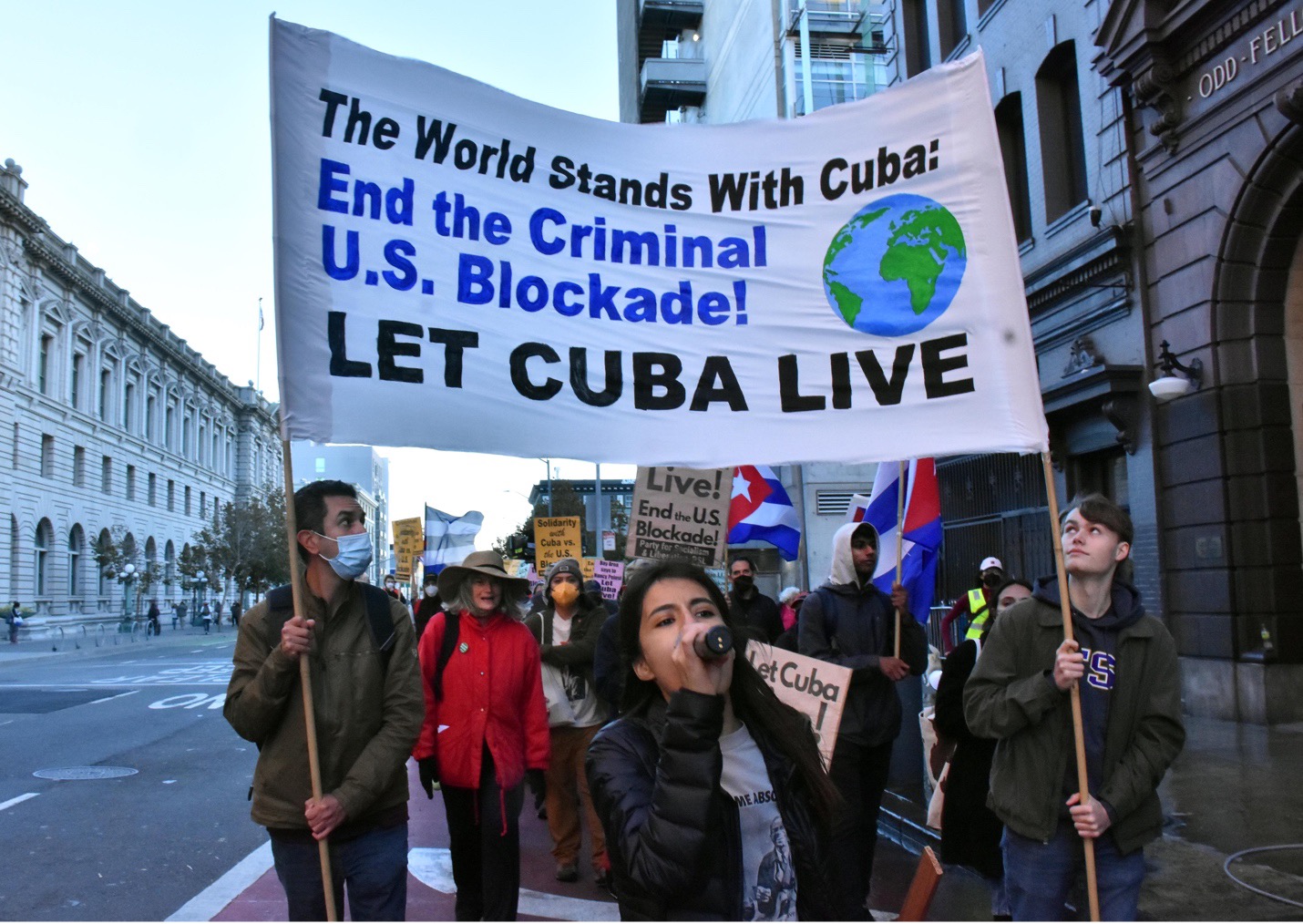 picture of protestors walking down a street with banner that reads: "The world stands with Cuba End the criminal U.S. blockade! LET CUBA LIVE Photo by: Bill Hackwell